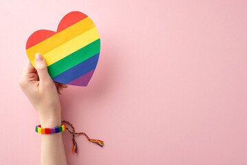 LGBT History Month theme: first person top view photo of young woman's hand in symbolic bracelet holding rainbow heart card on pastel pink background with space for text or advert