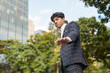 © Mdv Edwards - A stylish guy in a blue checkered suit in a serious face with a reaching his hand down gesture while standing outside, in the city. Sky, building and trees are shown in the background.