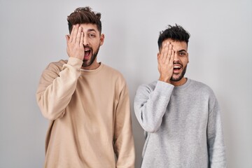 Young homosexual couple standing over white background covering one eye with hand, confident smile on face and surprise emotion.