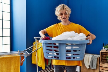 Poster - Young blond man smiling confident holding basket with clothes at laundry room