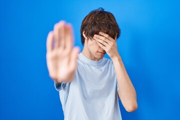 Poster - Hispanic young man standing over blue background covering eyes with hands and doing stop gesture with sad and fear expression. embarrassed and negative concept.