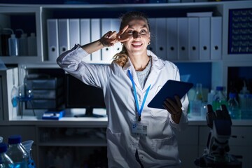 Poster - Beautiful blonde woman working at scientist laboratory late at night doing peace symbol with fingers over face, smiling cheerful showing victory