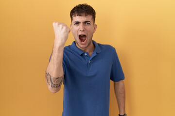 Sticker - Young hispanic man standing over yellow background angry and mad raising fist frustrated and furious while shouting with anger. rage and aggressive concept.