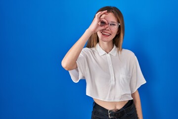 Poster - Beautiful woman standing over blue background doing ok gesture with hand smiling, eye looking through fingers with happy face.