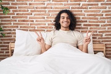 Wall Mural - Young latin man doing victory gesture sitting on bed at bedroom