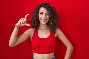 Sticker - Hispanic woman with curly hair standing over red background smiling and confident gesturing with hand doing small size sign with fingers looking and the camera. measure concept.