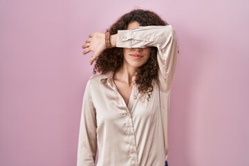 Sticker - Hispanic woman with curly hair standing over pink background covering eyes with arm, looking serious and sad. sightless, hiding and rejection concept