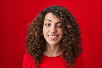 Sticker - Hispanic woman with curly hair standing over red background winking looking at the camera with sexy expression, cheerful and happy face.