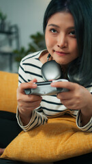 Excited beautiful Asian woman using joystick wireless controller enjoy playing fighting console video game to win highest victory. Girl Gamer sit on sofa in modern living room at home