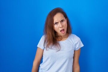 Wall Mural - Brunette woman standing over blue background in shock face, looking skeptical and sarcastic, surprised with open mouth