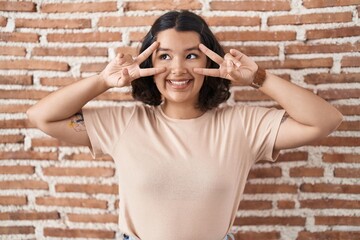 Canvas Print - Young hispanic woman standing over bricks wall doing peace symbol with fingers over face, smiling cheerful showing victory