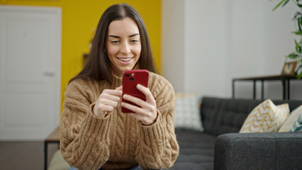 Wall Mural - Young beautiful hispanic woman using smartphone sitting on sofa at home