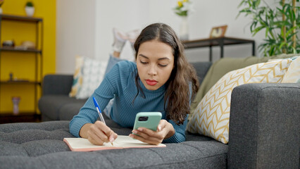 Poster - Young beautiful hispanic woman using smartphone writing on notebook lying on sofa at home