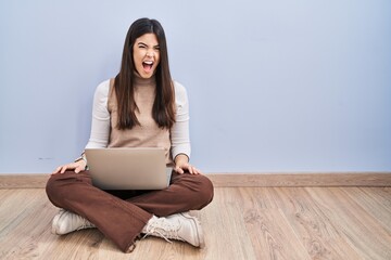 Sticker - Young brunette woman working using computer laptop sitting on the floor angry and mad screaming frustrated and furious, shouting with anger. rage and aggressive concept.