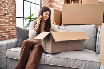 Sticker - Young beautiful hispanic woman talking on smartphone unpacking cardboard box at new home