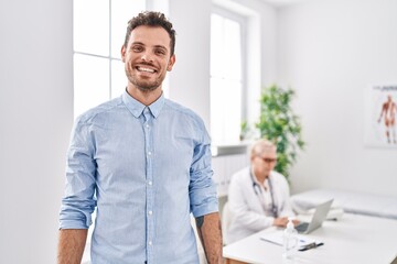 Sticker - Hispanic man at doctor clinic looking positive and happy standing and smiling with a confident smile showing teeth