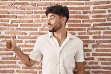 Poster - Arab man with beard standing over bricks wall background looking proud, smiling doing thumbs up gesture to the side