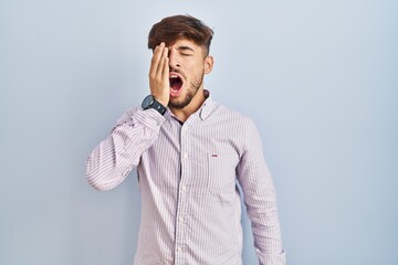 Poster - Arab man with beard standing over blue background yawning tired covering half face, eye and mouth with hand. face hurts in pain.