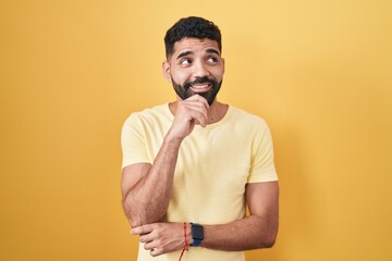 Wall Mural - Hispanic man with beard standing over yellow background with hand on chin thinking about question, pensive expression. smiling and thoughtful face. doubt concept.