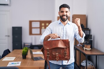 Wall Mural - Hispanic man with beard working at the office holding briefcase screaming proud, celebrating victory and success very excited with raised arm