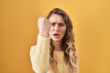 Wall Mural - Young caucasian woman standing over yellow background angry and mad raising fist frustrated and furious while shouting with anger. rage and aggressive concept.