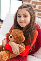 Sticker - Adorable hispanic girl hugging teddy bear sitting on bed at bedroom