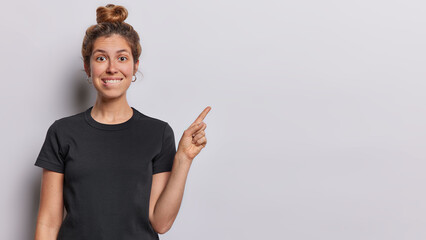 waist up shot of cheerful beautiful woman bites lips and points index finger at copy space for your 