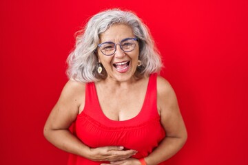 Wall Mural - Middle age woman with grey hair standing over red background smiling and laughing hard out loud because funny crazy joke with hands on body.