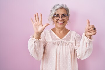 Canvas Print - Middle age woman with grey hair standing over pink background showing and pointing up with fingers number six while smiling confident and happy.