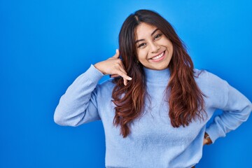 Sticker - Hispanic young woman standing over blue background smiling doing phone gesture with hand and fingers like talking on the telephone. communicating concepts.