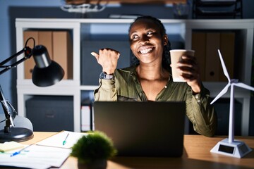 Canvas Print - African woman working using computer laptop at night smiling with happy face looking and pointing to the side with thumb up.