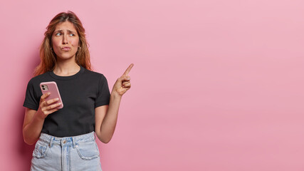 Wall Mural - Horizontal shot of displeased European woman purses lips points index finger aside on blank space tells something unpleasant wears casual black t shirt and jeans isolated over pink background