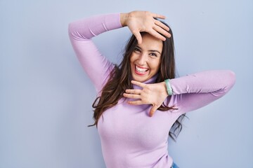 Poster - Young brunette woman standing over blue background smiling cheerful playing peek a boo with hands showing face. surprised and exited