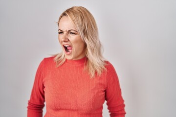 Wall Mural - Blonde woman standing over isolated background angry and mad screaming frustrated and furious, shouting with anger. rage and aggressive concept.