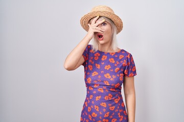 Poster - Young caucasian woman wearing flowers dress and summer hat peeking in shock covering face and eyes with hand, looking through fingers with embarrassed expression.