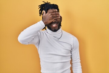 Sticker - African man with dreadlocks wearing turtleneck sweater over yellow background smiling and laughing with hand on face covering eyes for surprise. blind concept.