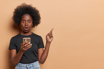 horizontal shot of impressed black woman with bushy hair holds mobile phone reacts to something asto