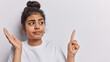 Horizontal shot of pretty Indian girl keeps palm raised up feels hesitant points index finger aside on copy space for your advertising content isolated over white background recommends to look there