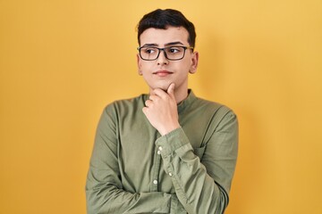 Poster - Non binary person standing over yellow background looking confident at the camera with smile with crossed arms and hand raised on chin. thinking positive.