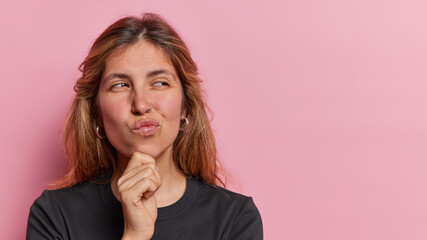 Wall Mural - Beautiful European woman purses lips keeps hand on chin concentrated aside ponders on decision dressed in black t shirt has thoughtful face isolated over pink background copy space for your promotion