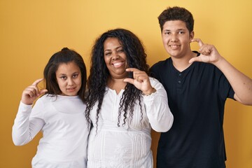 Sticker - Family of mother, daughter and son standing over yellow background smiling and confident gesturing with hand doing small size sign with fingers looking and the camera. measure concept.