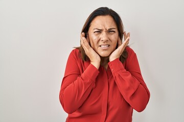 Poster - Hispanic mature woman standing over white background covering ears with fingers with annoyed expression for the noise of loud music. deaf concept.