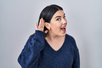 Canvas Print - Young brunette woman standing over isolated background smiling with hand over ear listening an hearing to rumor or gossip. deafness concept.