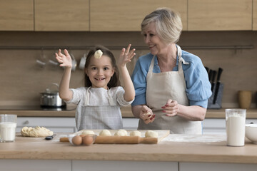 Attractive retired granny teach cute funny granddaughter to cook, prepare buns or cookies, make handmade pastries, happy child throwing dough looks playful white cooking with loving grandma in kitchen