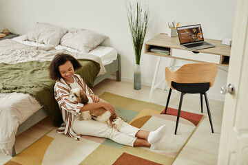 Full length portrait of happy young woman sitting on floor at home and cuddling with pet dog in cozy interior, copy space