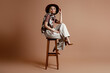 © gstockstudio - Fashionable young woman adjusting her elegant hat while sitting on a chair against brown background