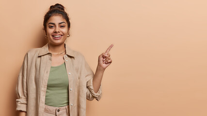 Studio shot of pretty young Indian girl points index finger at mock up copy space demonstrates new offer or great opportunities has happy smile on face wears shirt isolated over brown background
