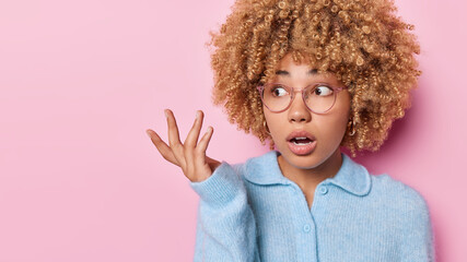 Wall Mural - Attractive puzzled curly haired woman feels doubt and hesitation shrugs shoulders and looks clueless aside dressed in casual blue jumper isolated over pink background blank copy space aside.