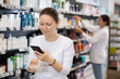 © JackF - Adult woman in casual clothes looking for cosmetic product from photo on internet on smartphone in pharmacy..