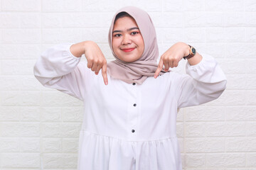 Positive Asian muslim woman pointing down with two fingers, demonstrating free place for your design or advertisement, standing over white background